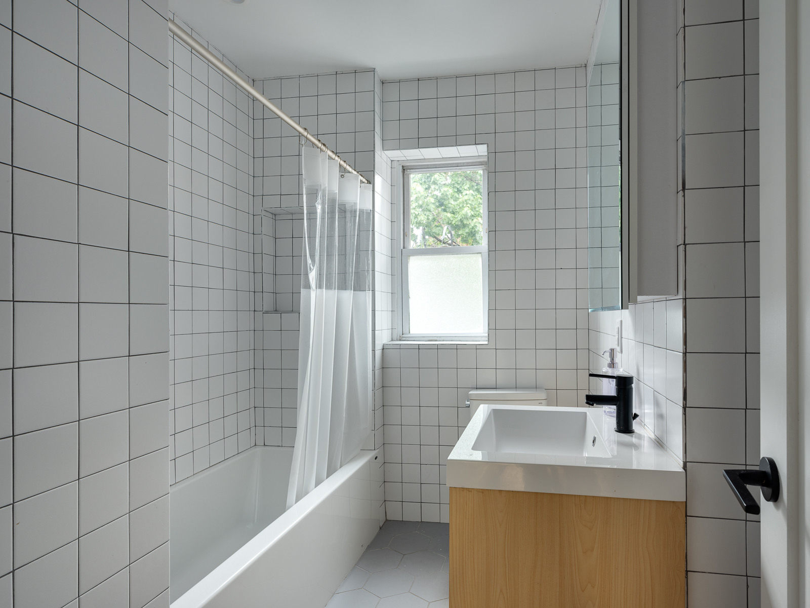White square-tiled bathroom with soaking tub, oak vanity and a window onto trees